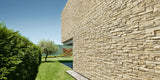 Modern home exterior featuring beige stacked stone cladding with a natural finish, set in a landscaped garden with manicured lawn and trees under a clear blue sky